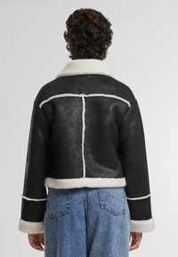 Veste en shearling noir avec un col et des bordures en laine blanche, design court, finition texturée et détails de couture visibles au dos.
