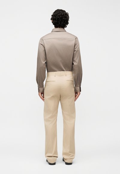 Άνδρας με σγουρά σκουρόχρωμα μαλλιά, που φοράει καφέ πουκαμισο με μακριά μανίκια, κρυμμένο μέσα σε μπεζ παντελόνια, στέκεται γυρισμένος με την πλάτη σε λευκό φόντο.