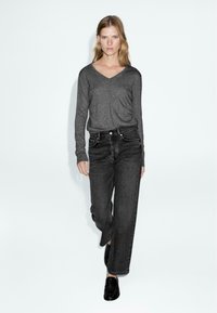 Maglietta grigia a maniche lunghe con scollo a V abbinata a jeans neri a vita alta. Il modello indossa mocassini neri; l'immagine presenta uno sfondo bianco semplice.