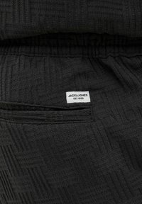 Crni, teksturirani joggeri od tkanine s malom bijelom etiketom na kojoj piše "JACK & JONES EST. 1990" te detaljom džepova. Vidljivi elastični pojas.