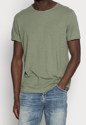 Dunkelhäutige Person, die ein olivgrünes kurzärmeliges T-Shirt und hellblaue, ausgewaschene Jeans trägt, mit einer Hand in der Tasche.