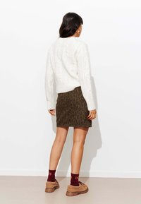 Pull en maille blanche, jupe à imprimé léopard vert foncé, chaussettes bordeaux avec un numéro, et chaussures marron. Look simple, texturé et à motifs.