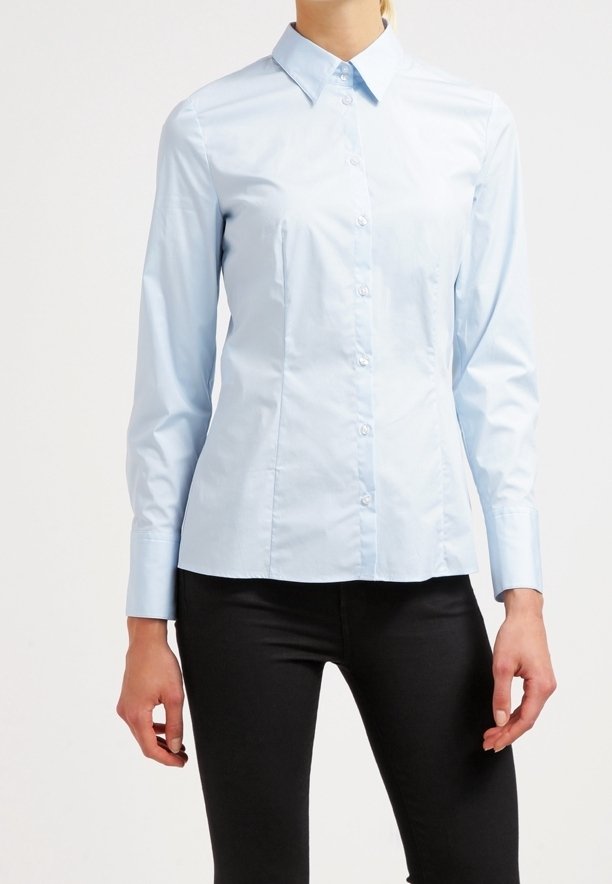 Femme portant une chemise bleu clair à manches longues boutonnée et un pantalon noir, debout devant un fond clair uni.