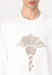 Sudadera blanca con un diseño recortado en forma de flor con acentos metálicos. Tejido texturizado con escote redondo.