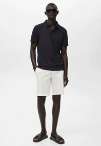 Svart polo skjorta, vita shorts, svarta slidesandaler. Modell med solglasögon, står rakt med ena handen i fickan.