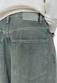 Jeans de denim verde pal, care prezintă două buzunare pe spate, o textură netedă și o etichetă rectangulară pentru talie într-o culoare contrastantă.