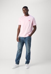 Lichtroze katoen t-shirt met een rode hartafbeelding op de borst, gecombineerd met vervaagde blauwe denim jeans en witte sneakers.