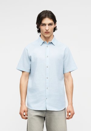 Młody mężczyzna o średniej długości ciemnych włosach, ubrany w jasnoniebieską koszulę na krótki rękaw zapinaną na guziki oraz jasnoszare spodnie, stojący prosto.