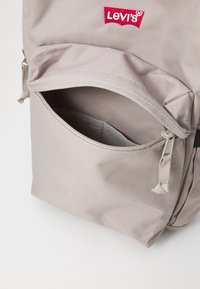 Světle šedý batoh s texturovanou látkou, přední kapsa na zip a červené logo Levi's. Nastavitelné popruhy a minimalistický design.