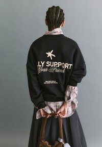 Čierny oversize sveter s bielym nápisom "ALLY SUPPORT Your Friends". Nosený cez károvanú košeľu. Má rebrované manžety a pásik.