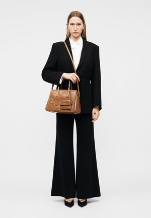 Donna con completo nero, camicia bianca, tacchi neri, che tiene una borsa a tracolla in pelle marrone, in piedi davanti a uno sfondo semplice.