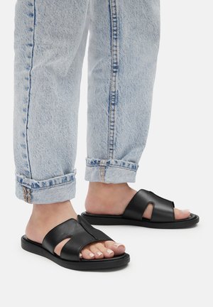 Czarne skórzane klapki z otwartym palcem, z wyciętym wzorem; zestawione z podwiniętymi jasnoniebieskimi dżinsami. Płaska podeszwa, minimalistyczny styl.