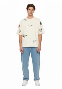 Off-white oversized t-shirt med sorte grafiske tryk, herunder tekst og symboler. Parret med lyseblå denim jeans og hvide sneakers.