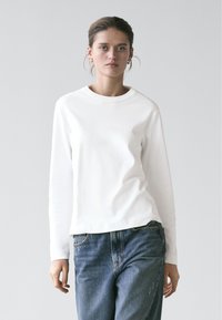 Young woman with tied-back hair wears white long-sleeve shirt and blue jeans, standing against a plain light gray background.