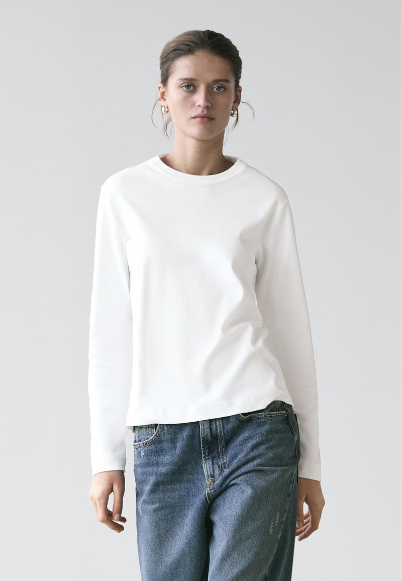 Young woman with tied-back hair wears white long-sleeve shirt and blue jeans, standing against a plain light gray background.