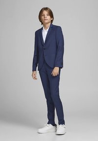 Marineblå jakkesæt med en figursyet blazer og matchende bukser, kombineret med en hvid skjorte og hvide sneakers. Glat stof, to-knaps lukning.