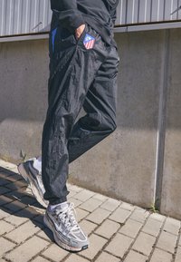 Black, textured athletic pants with blue accents and a team logo on the side, paired with gray Nike running shoes on a paved surface.