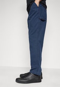 Pantaloni in cotone blu navy con taglio dritto, dotati di tasche laterali e una cucitura a contrasto. Abbinati a sneakers sportive nere.