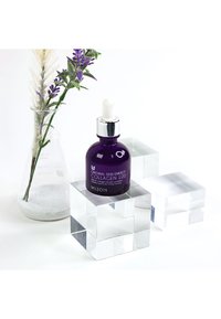 Lila glasserumflaska med pipett, märkt "Collagen 100," placerad på transparenta akrylblock. I bakgrunden en vas med torkade blommor.