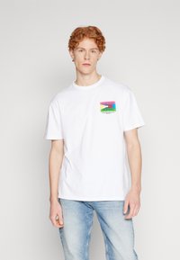 Camiseta de algodón blanca con cuello redondo, que presenta un estampado gráfico colorido en la parte izquierda del pecho. Mangas de corte recto, ajuste relajado.