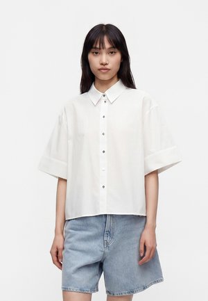 Femme portant une chemise blanche ample à manches courtes avec boutons et un short en jean bleu clair sur un fond blanc uni.