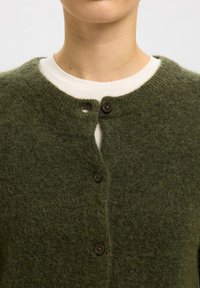 Primer plano de una persona que lleva una rebeca abotonada de color verde oscuro y peluda sobre una camiseta blanca de cuello redondo, mostrando el cuello y la parte superior del pecho.