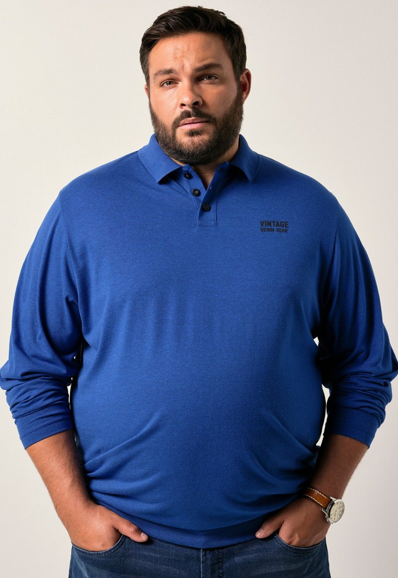 Mies, jolla on parta, pukeutuneena siniseen pitkähihapaitaan, jossa lukee "Vintage Denim Gear", kädet taskuissa ja rannekello vasemmassa ranteessa.