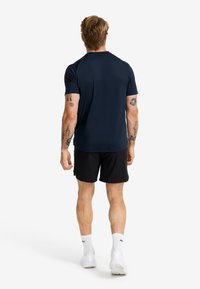 Marinblått kortärmat träningströja, svarta shorts och vita sneakers. Slät tyg, avslappnad passform, med synliga tatueringar på armarna.