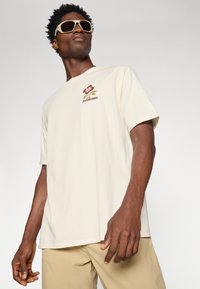 Patagonia krém színű pamut póló rövid ujjal, virágmintával és logóval a mellrészén. Barna nadrággal és oversized napszemüveggel viselve.