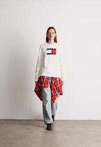 Λευκή μπλούζα με τύπωμα λογότυπου, συνδυασμένη με φαρδιά ανοιχτό μπλε τζιν και ένα κόκκινο καρό πουκάμισο δεμένο γύρω από τη μέση. Άνετα υποδήματα ολοκληρώνουν το σύνολο.