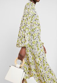 Robe à imprimé floral avec des accents verts et noirs, manches longues bouffantes, tissu fluide. Sac à main blanc avec poignée en bois.