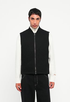 Black sleeveless vest with a high collar, front zipper, and side pockets, worn over a white sweater. The fabric has a smooth texture.