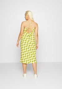 Vestido midi amarelo com estampado floral e padrões de folhas verdes, apresentando um design de costas cruzadas e detalhe de laço, combinado com sapatos brancos.