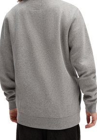 Osoba v jednoduchém šedém dlouhém svetru viděná ze zad s černými kalhotami, stojící s uvolněnýma rukama podél těla.