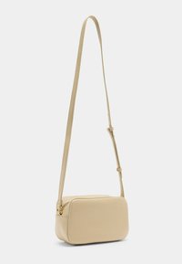 Väike ristküliku kujuline beežist crossbody kott, millel on kareda tekstuuriga pind ja reguleeritav rihm, ripub valge tausta ees.