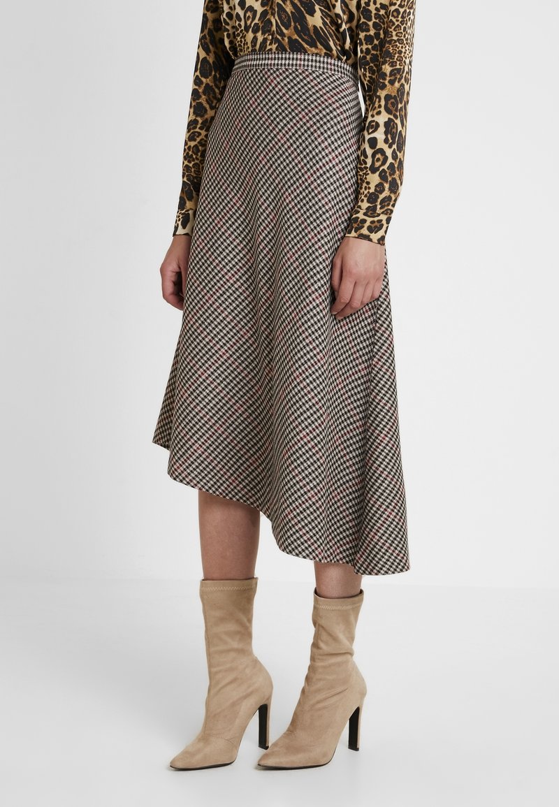 Femme portant une jupe à carreaux longueur genoux, un haut à manches longues imprimé léopard, et des bottines en daim beige à talons, debout sur un sol blanc.