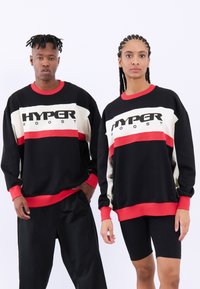 Čierna mikina s červenými a krémovými pruhmi a logom "HYPER boost." Pružné manžety a lem. Model má na sebe čierne šortky; textúrovaná tkanina.