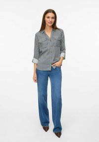 Chemise rayée noire et blanche avec deux poches poitrine, fermeture à boutons à l'avant et manches retroussées. Associée à un jean large bleu et des talons noirs.