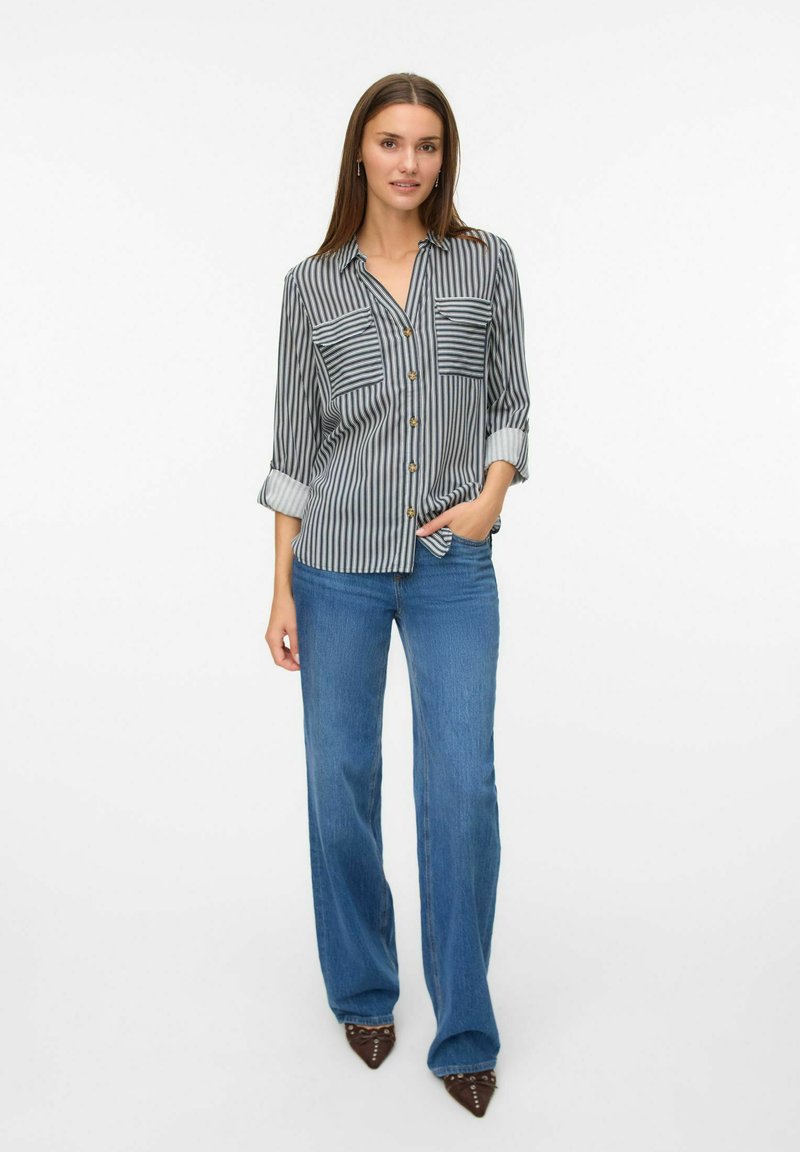 Chemise rayée noire et blanche avec deux poches poitrine, fermeture à boutons à l'avant et manches retroussées. Associée à un jean large bleu et des talons noirs.