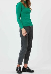 Grön långärmad topp, grå tapered jeans och glänsande svarta loafers. Outfiten har en figurnära design och texturerade strumpor.