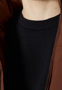 Giacca marrone con zip su un maglione nero. La giacca ha una texture morbida con una cerniera liscia, mentre il maglione presenta un collo a coste.