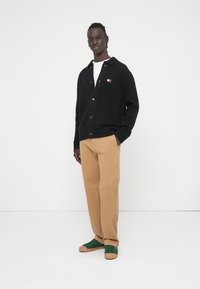 Μαύρο ζακέτα με γιακά, κουμπιά μπροστά και πλεκτές μανσέτες, συνδυασμένο με μπεζ παντελόνι ίσιας γραμμής και πράσινα αθλητικά παπούτσια.