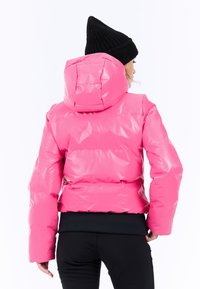 Lyserød puffjakke med en blank finish, med hætte og quiltet design. En sort ribstrikket hue bæres, med en tætsiddende mørk top under.