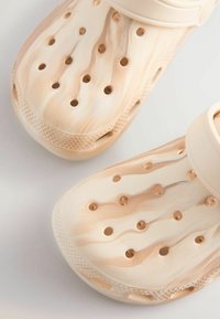 Beige träskor med marmorering, utrustade med flera ventilationshål och ett band över toppen för säker passform.