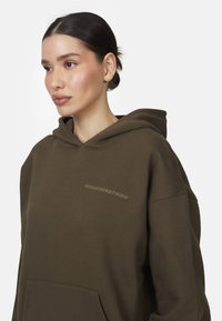 Haut à capuche vert olive avec une grande poche et un cordon de serrage. Présente un logo "GOODFORNOTHING" sur la poitrine. Tissu doux et texturé.