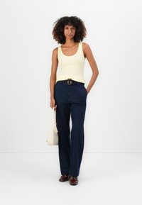 Top amarillo sin mangas combinado con pantalones oscuros de tiro alto y pierna ancha, con costuras detalladas y un cinturón con hebilla dorada. Accesorio: bolso pequeño blanco.