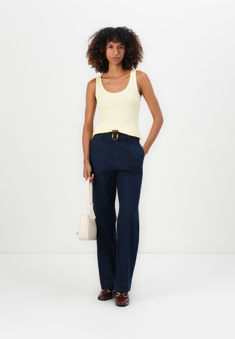 Top amarillo sin mangas combinado con pantalones oscuros de tiro alto y pierna ancha, con costuras detalladas y un cinturón con hebilla dorada. Accesorio: bolso pequeño blanco.