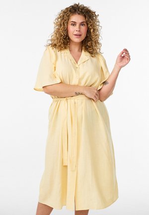 Žena s kovrčavom kosom nosi svijetložutu haljinu s pojasom i kopčama, pozira s jednim savijenim rukom, a drugom blago podignutom.