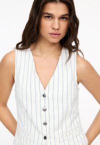 Witte mouwloze vest met blauwe verticale strepen, V-hals en vijf zilveren knopen. Beschikt over twee voorzakken en een textuurstof.