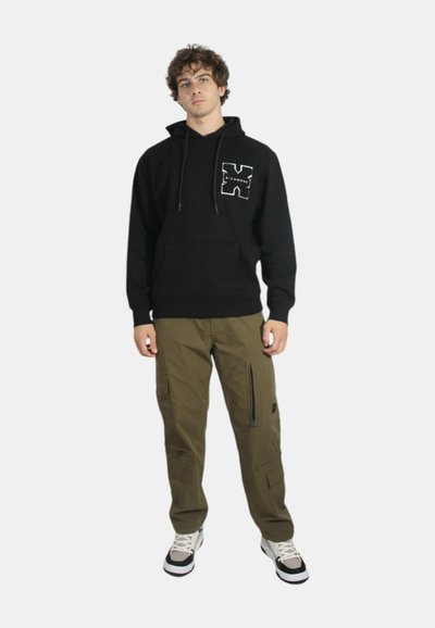 Felpa nera con tasca frontale e logo bianco, abbinata a pantaloni cargo verde oliva con tasche laterali e dettagli neri. Sneakers bianche.